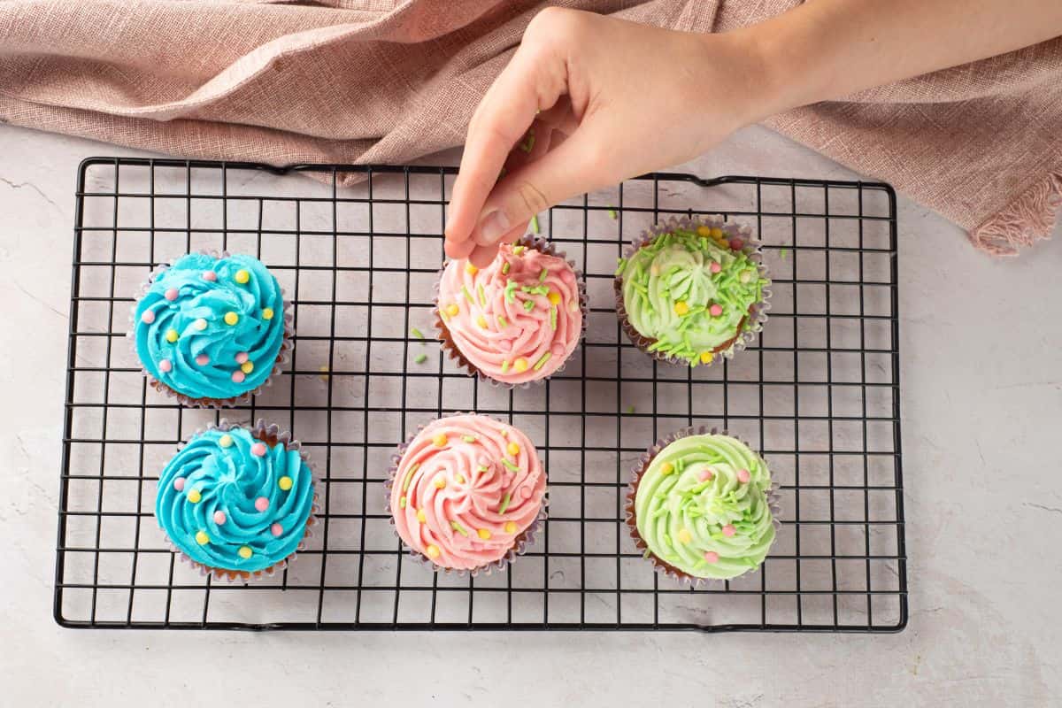 Delicious decorated cupcakes in pink, blue, and green with colorful sprinkles, cooling on a wire rack.